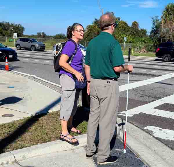 Street view image of Jen Graham COMS and Jeff Thompson PE standing at the corner facing the street.  Jeff’s back is to the camera and he is holding a white cane parallel to his body and wearing a set of vision simulators.  Jen stands to his left in the grass.